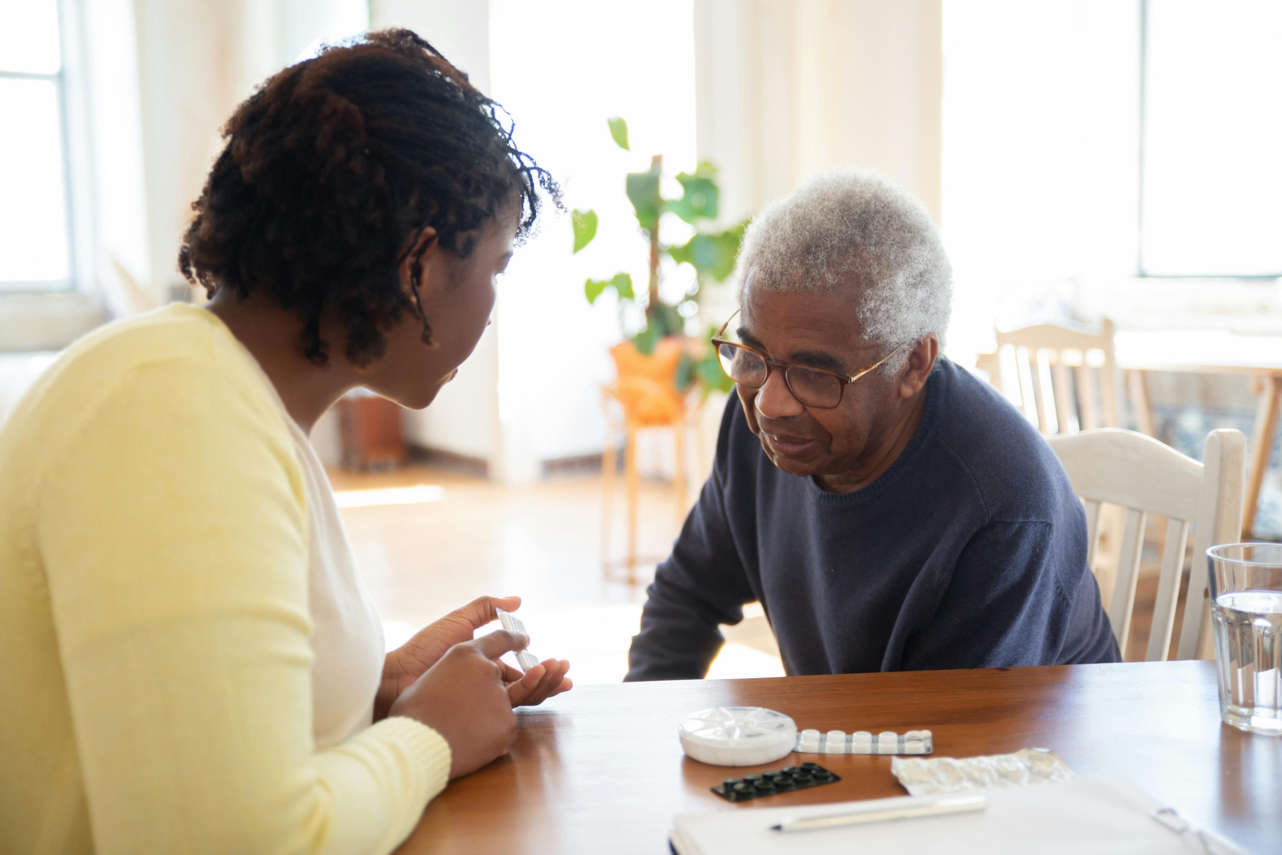 A woman and an elderly man discuss medication at a wooden table in a bright room, highlighting care.